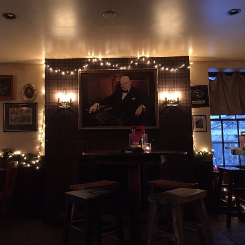 A cozy pub interior with a large portrait of a man, surrounded by string lights and wooden stools.