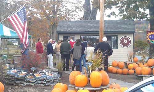 A group of people stands in line outside a small shop surrounded by pumpkins and autumn foliage. An American flag is visible.
