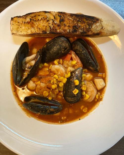 A bowl of seafood stew with shrimp, mussels, corn, and a toasted piece of bread on the side.