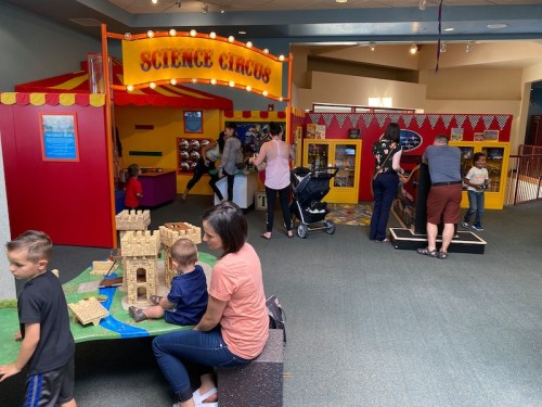 Children and adults explore a colorful science-themed play area with interactive exhibits and a castle play structure.