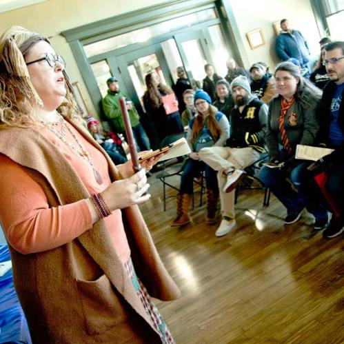 A woman speaks to an engaged audience seated in a room, with people listening attentively in the background.