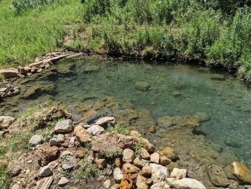 A clear, shallow pond surrounded by green grass and rocky edges, reflecting the blue sky above.