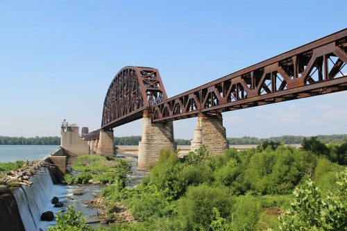 A large metal bridge spans a river, supported by stone pillars, with greenery and a dam visible in the foreground.