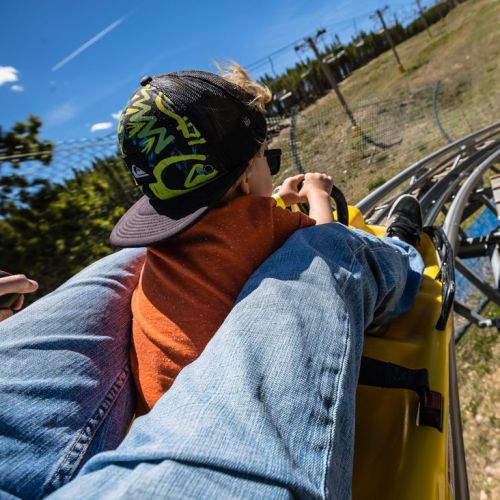 A child wearing a cap enjoys a roller coaster ride, gripping the safety bar with excitement against a blue sky.