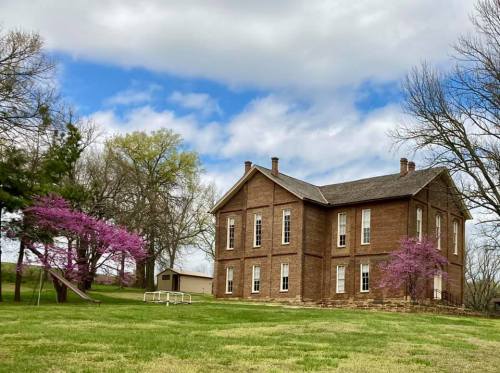 A historic brick building surrounded by green grass and blooming pink trees under a partly cloudy sky.