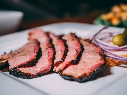 Sliced smoked brisket on a white plate, accompanied by red onion and a pickle.