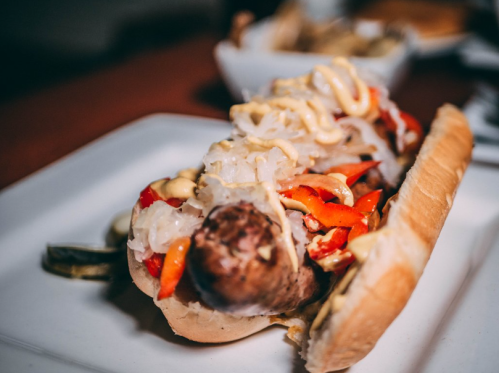 A close-up of a loaded hot dog topped with sauerkraut, mustard, and colorful peppers in a soft bun.