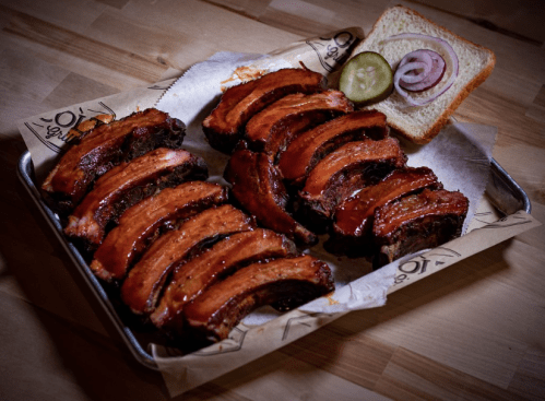 A platter of glazed ribs with a slice of bread, pickles, and red onion on a wooden table.
