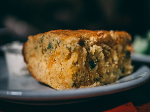 A slice of cornbread on a plate, with a small cup of butter or cream beside it.