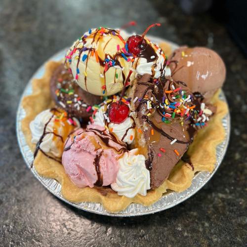 A colorful ice cream sundae in a pie crust, topped with whipped cream, cherries, sprinkles, and chocolate sauce.
