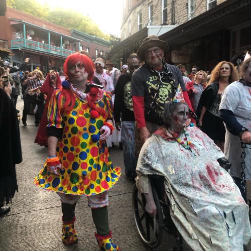 A colorful clown and a zombie in a wheelchair lead a group of costumed people in a festive street scene.