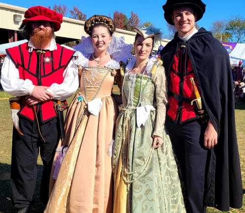 Four people in historical costumes stand together outdoors, smiling, with colorful tents in the background.