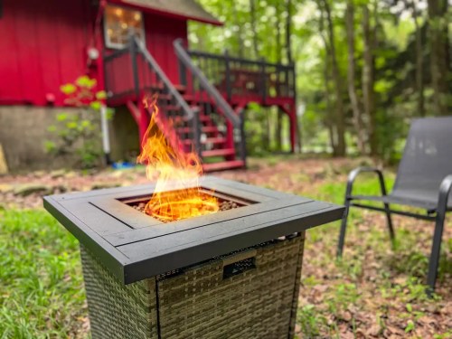A fire pit with flames in a wooded area, with a red house and a chair visible in the background.