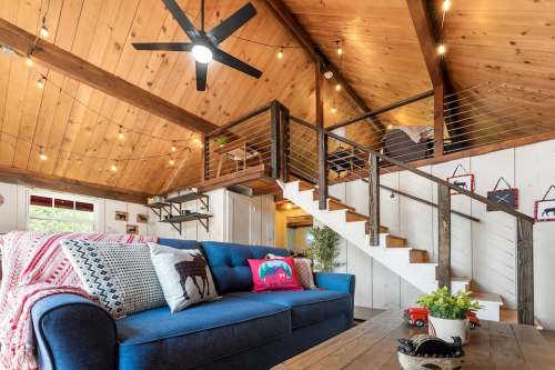 Cozy living room with a blue sofa, wooden ceiling, and a staircase leading to a loft area, adorned with string lights.