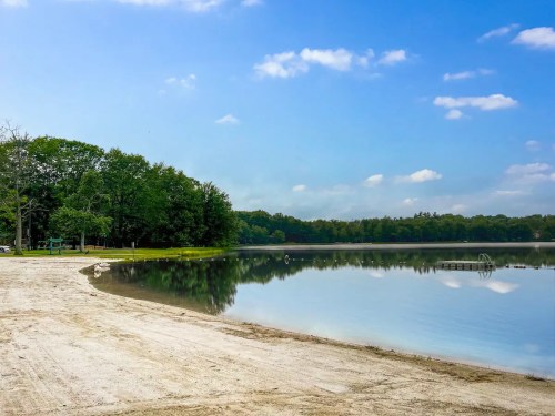 A serene lake surrounded by trees, with a sandy shore and a clear blue sky reflecting on the water's surface.