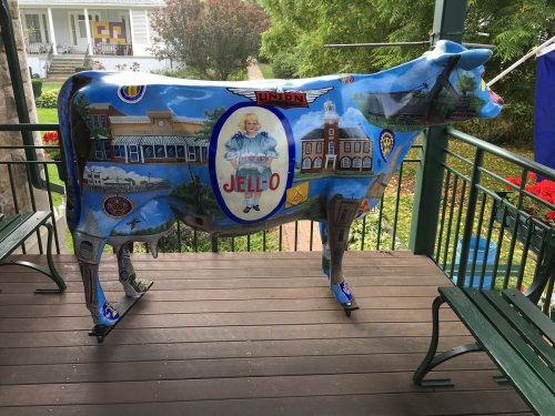 A colorful cow statue decorated with Jell-O themes and local landmarks, displayed on a wooden porch.