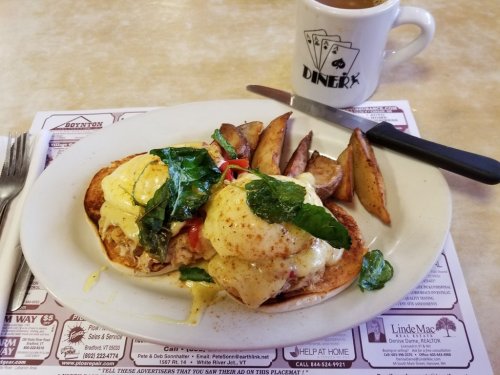 A plate of eggs Benedict with spinach and tomatoes, served with potato wedges and a coffee mug in the background.