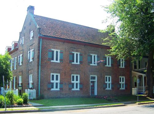 Historic brick building with a steep roof, white windows, and a green lawn, surrounded by trees and a picket fence.