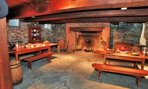 A rustic kitchen with wooden tables, stone floor, and a brick fireplace, featuring vintage cookware and utensils.