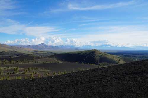 A panoramic view of rolling hills and mountains under a blue sky with scattered clouds, featuring dark volcanic soil.