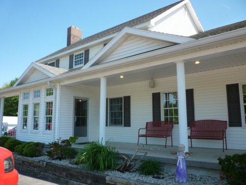 A white two-story house with a porch, red benches, and a garden, featuring a clear blue sky in the background.