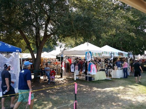 A bustling outdoor market with tents selling clothing and crafts, surrounded by trees and visitors exploring.