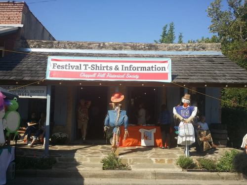 A storefront with a sign for "Festival T-Shirts & Information," featuring scarecrows and decorations outside.