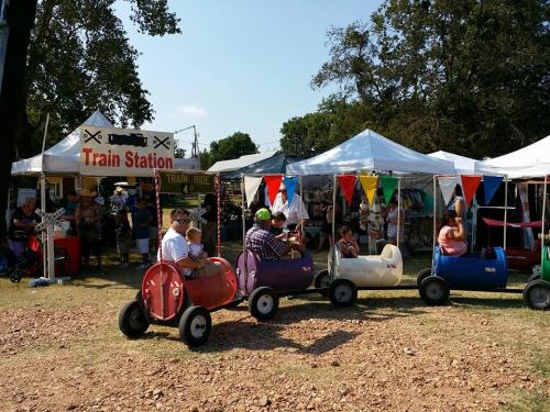 Children ride colorful mini trains at a fair, with a train station booth and tents in the background.