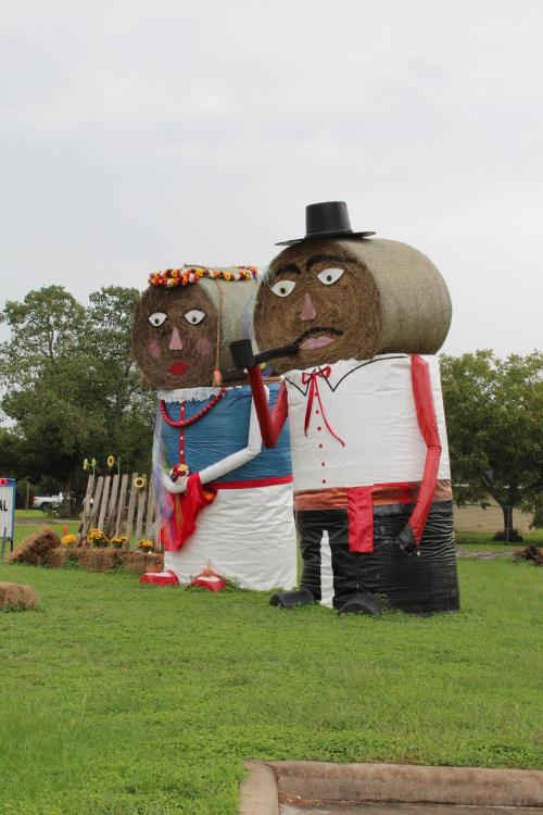 Two large hay bales dressed as a couple, with a hat and flowers, standing in a grassy field.
