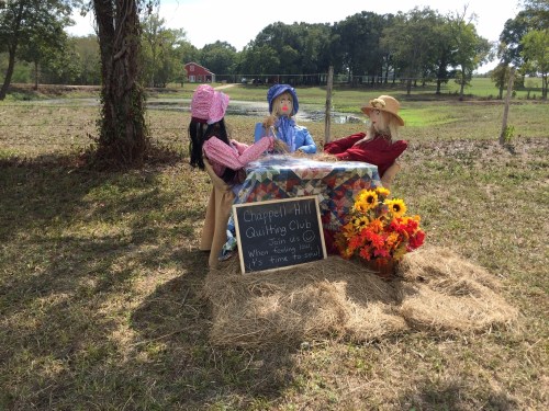 Scarecrows seated at a table with flowers, promoting the Chapel Hill Quilting Club in a rural setting.