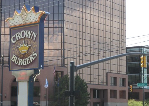 Sign for Crown Burgers in front of a modern building, with reflections in the glass and traffic signals nearby.