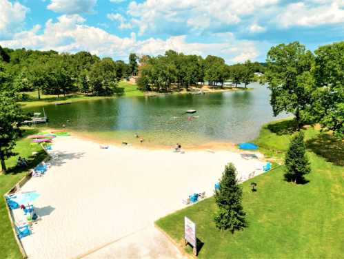 A serene lakeside scene with a sandy beach, green trees, and people enjoying the water on a sunny day.