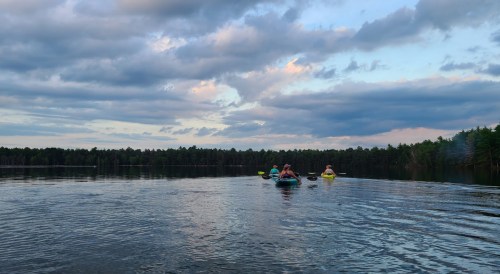 Two people kayak on a calm lake under a cloudy sky, surrounded by trees.