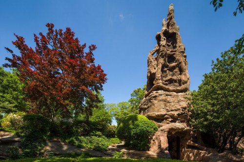 A tall rock formation surrounded by lush greenery and a vibrant red tree under a clear blue sky.