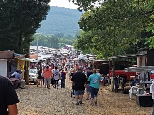 A bustling outdoor market with people walking along a gravel path, surrounded by stalls and greenery.