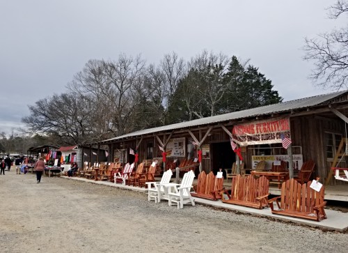 A rustic market with wooden furniture displays and shoppers browsing under a cloudy sky.