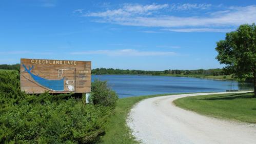 A sign for Czechland Lake beside a calm water body, with a gravel road and green trees under a blue sky.
