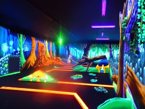 A vibrant, neon-lit mini-golf course featuring whimsical trees and colorful landscapes in a darkened room.
