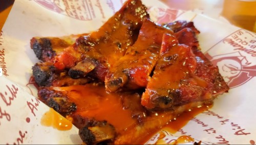 A plate of glazed barbecue ribs with a rich, sticky sauce, resting on a paper liner with printed text.