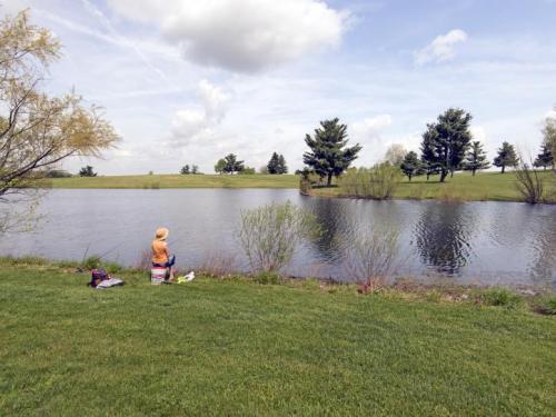 A child sits by a calm pond, surrounded by greenery and trees, under a partly cloudy sky.