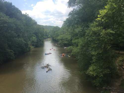 A serene river flows through a lush green landscape, with kayakers enjoying the water under a partly cloudy sky.