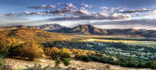 A panoramic view of rolling hills and mountains under a blue sky with scattered clouds, surrounded by lush greenery.