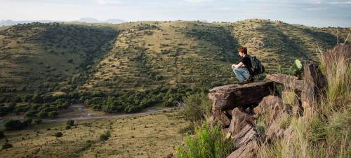 A person sits on a rocky ledge, overlooking a vast green landscape with rolling hills and scattered trees.
