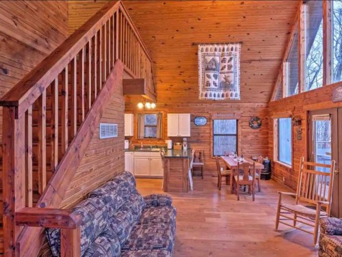 Cozy wooden cabin interior featuring a living area, kitchen, dining table, and staircase with large windows.