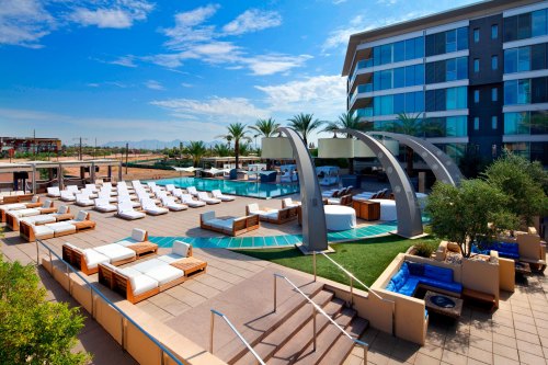 A modern hotel pool area with lounge chairs, palm trees, and a clear blue sky.