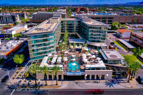 Aerial view of a modern hotel with a pool area, surrounded by palm trees and bustling cityscape in the background.