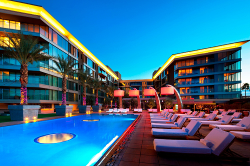 Modern hotel exterior at dusk, featuring a vibrant pool area with lounge chairs and palm trees.