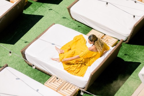 A woman in a flowing yellow dress relaxes on a sunbed surrounded by green grass and lounge chairs.