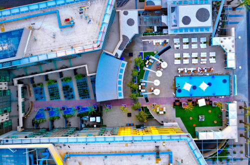 Aerial view of a modern rooftop with a pool, lounge area, and greenery, surrounded by buildings.