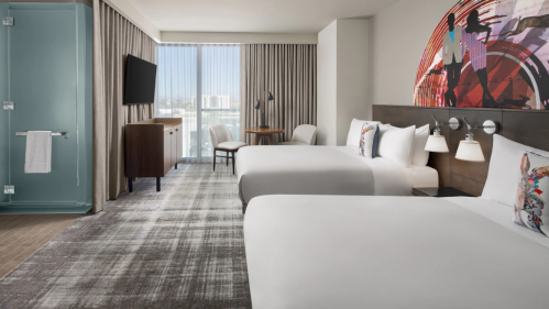 A modern hotel room featuring two beds, a TV, a desk, and large windows with city views.